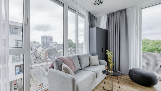 A modern living room with a grey sofa, a round black ottoman, and a small table with a vase. Large windows offer a view of the cityscape. - Limehome Mönchengladbach Fliethstraße (Moenchengladbach)
