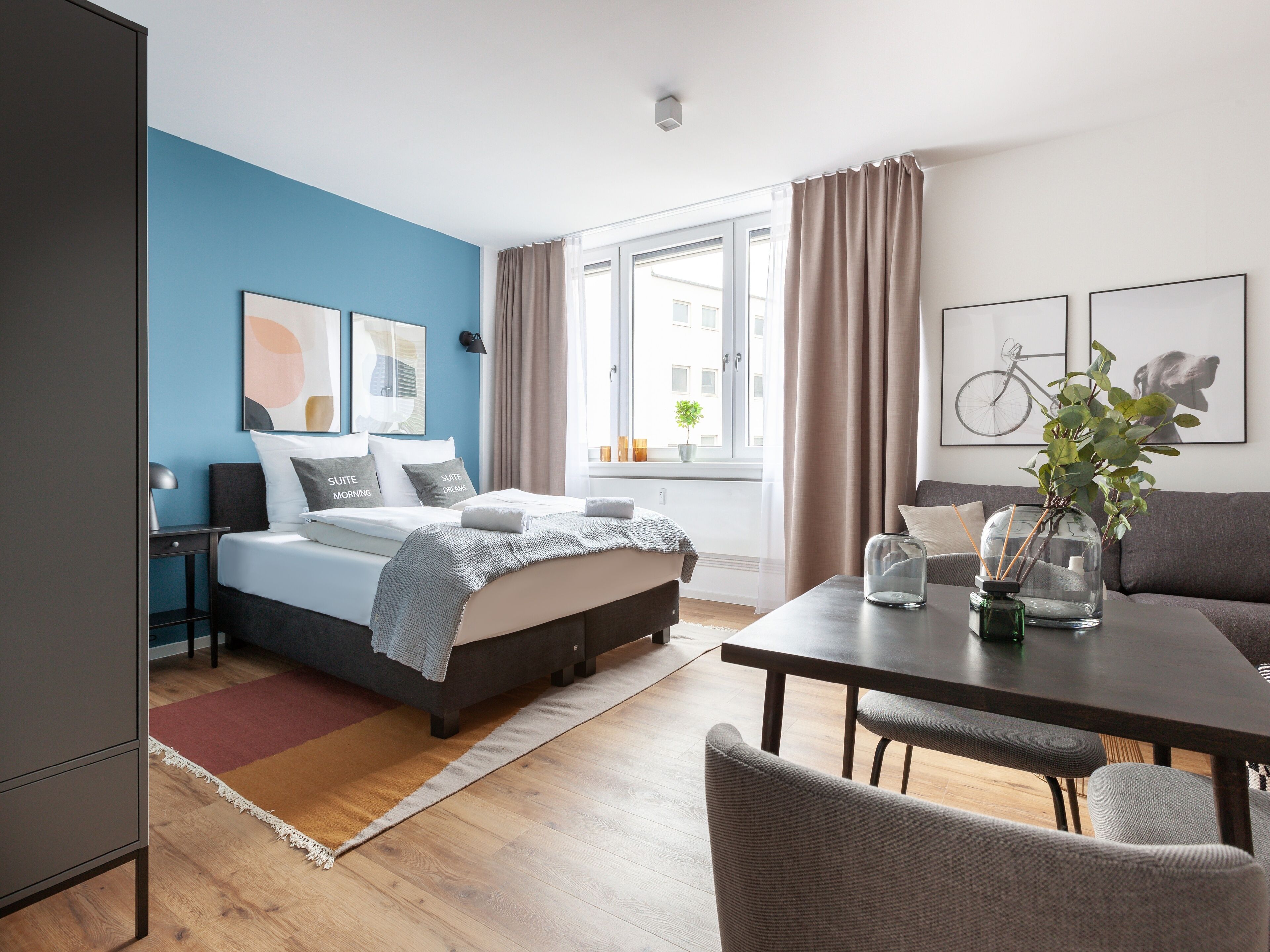 A modern hotel room with a bed, bedside table, and a dining area with a table and chairs.