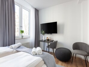 A modern hotel room with a bed, a desk, a chair, a round ottoman, a TV, and a window with curtains. - Limehome Bremen Hutfilterstraße (Bremen)
