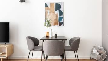 A modern living room with a flat-screen TV, a dining table with two chairs, a floor lamp, a wall-mounted abstract art piece, and a standing fan.