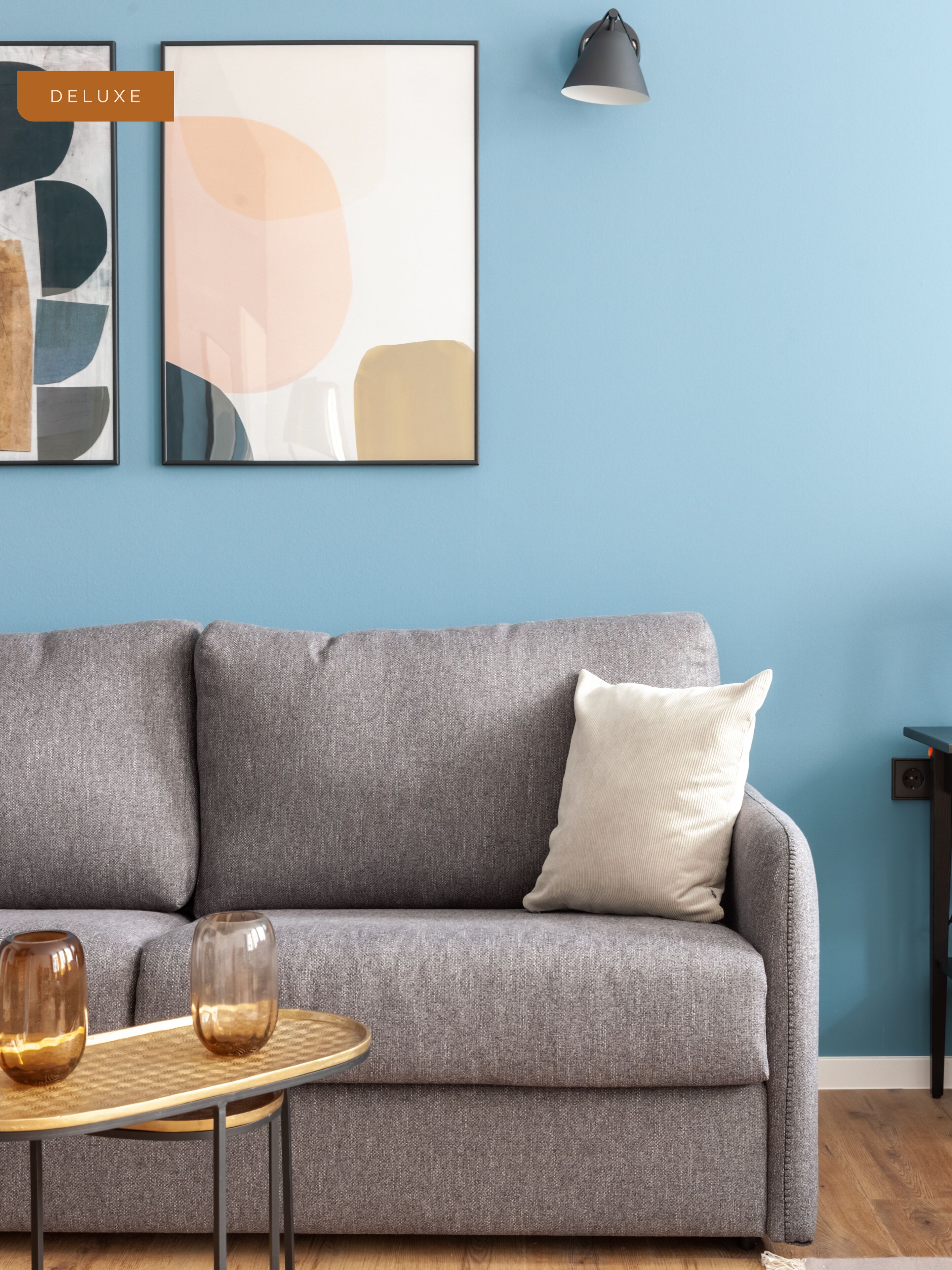Un sofá gris con un cojín blanco, una mesita redonda pequeña con dos jarrones de vidrio, y dos cuadros abstractos en la pared.