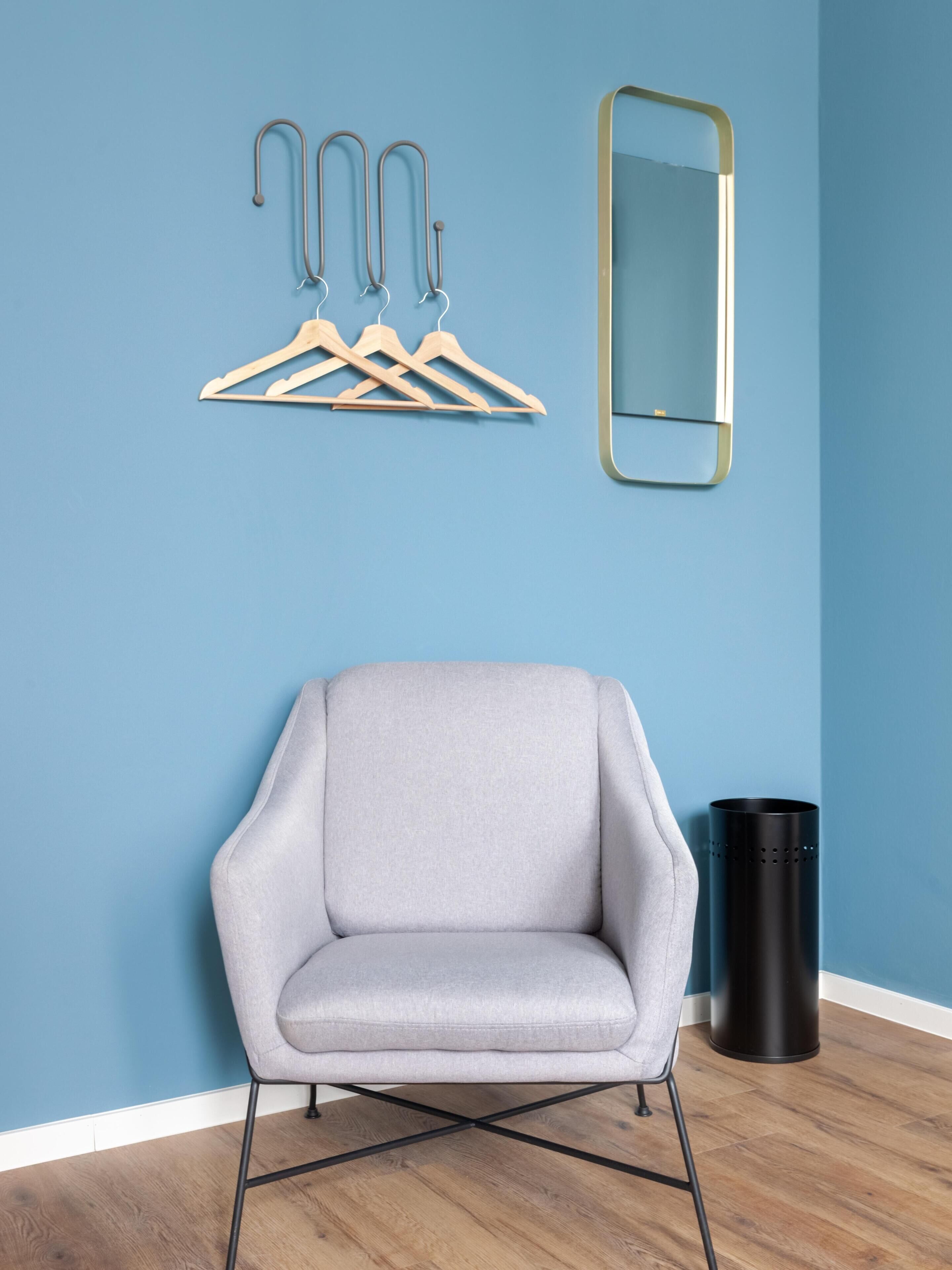 Ein Zimmer mit einem grau gepolsterten Sessel, hölzernen Kleiderhaken an der Wand, einem Spiegel und einem schwarzen Mülleimer.