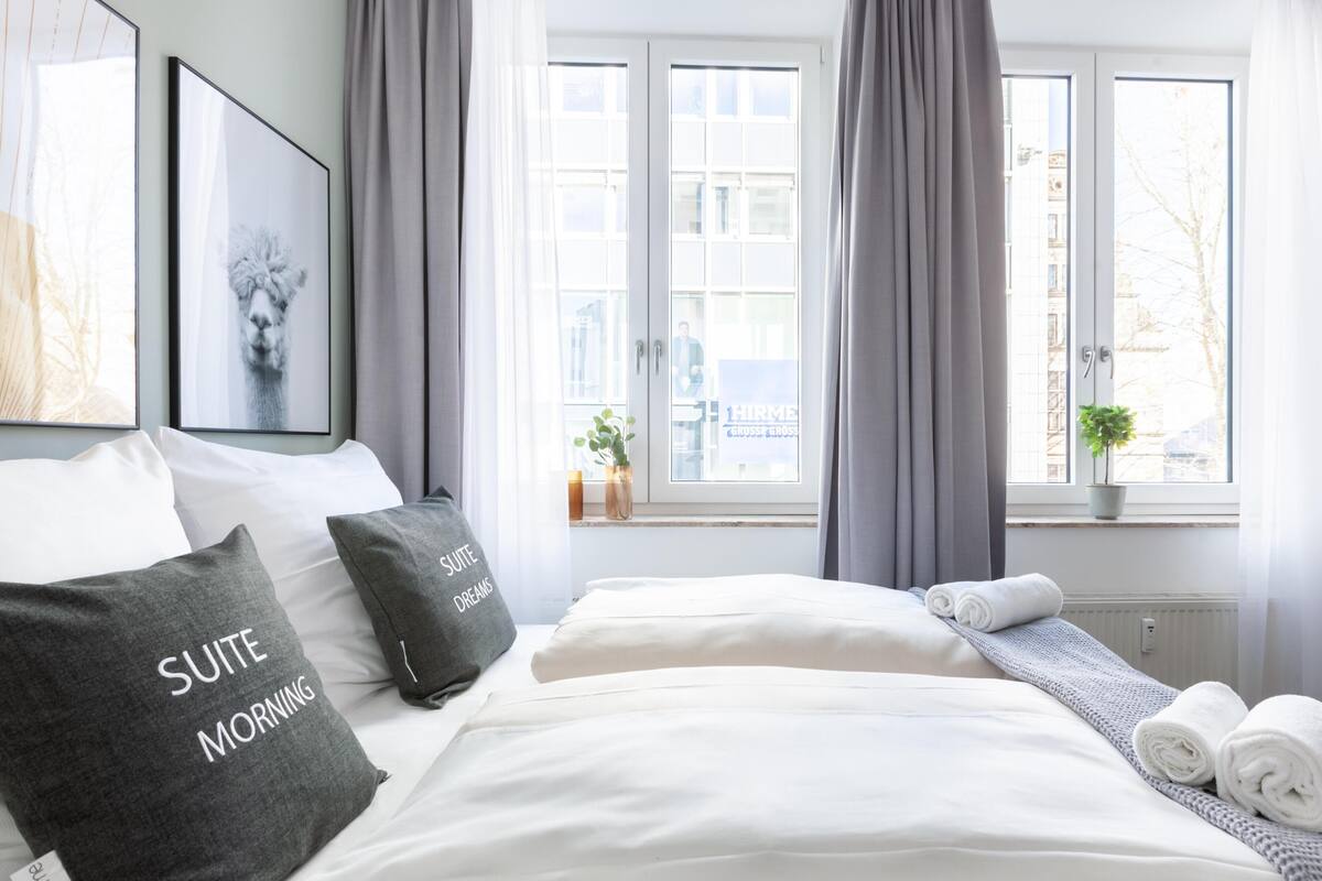 A hotel room with a neatly made bed, grey curtains, a window with a view of buildings, and a framed picture on the wall.