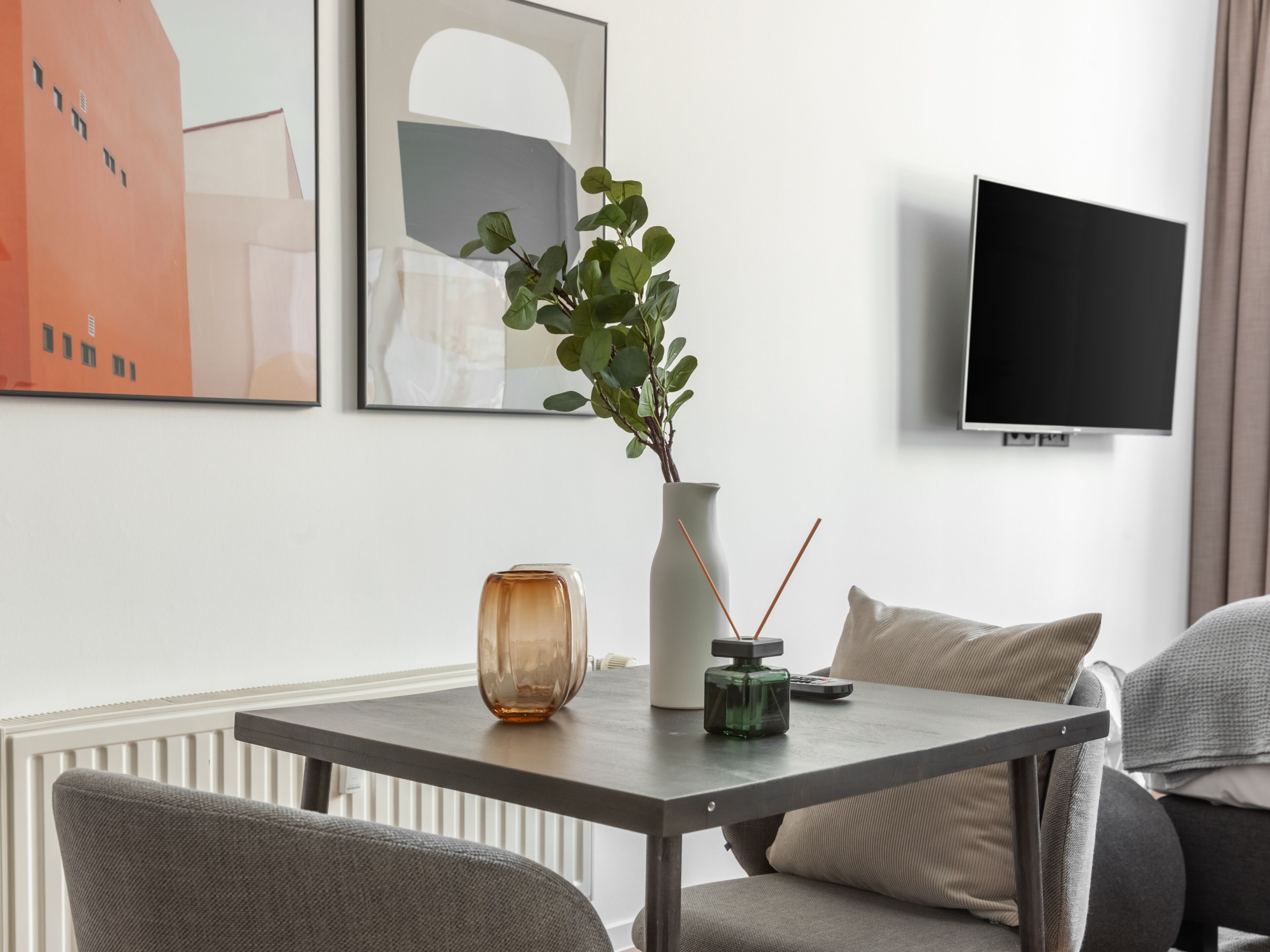 A modern hotel room with a dining table, a vase with a plant, a remote control, and a television on the wall.