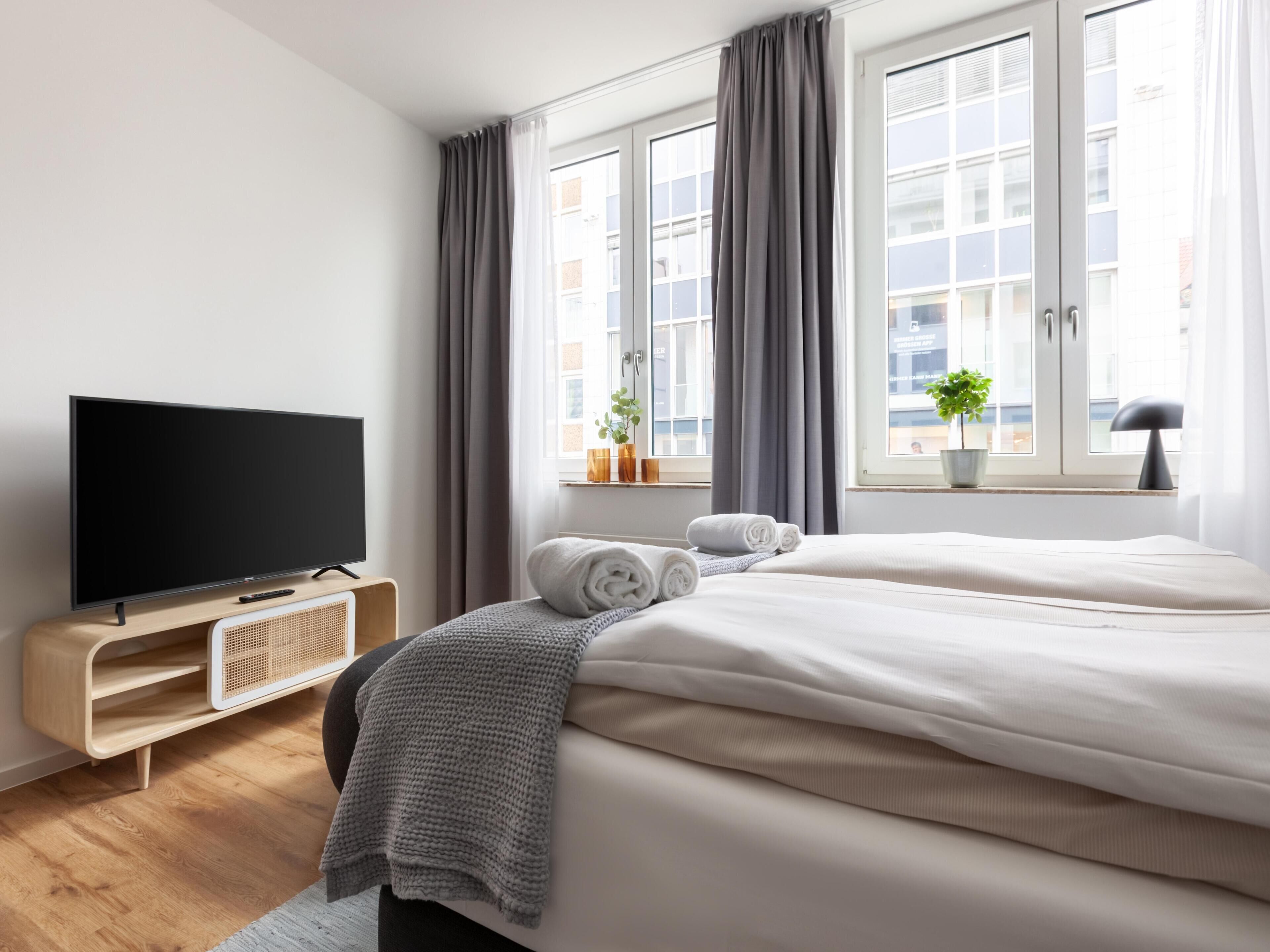 Ein modernes Hotelzimmer mit einem großen Bett, einem Flachbildfernseher, einem kleinen Tisch mit Pflanze und einem Fenster mit Vorhängen.