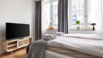 Ein modernes Hotelzimmer mit einem großen Bett, einem Flachbildfernseher, einem kleinen Tisch mit Pflanze und einem Fenster mit Vorhängen.