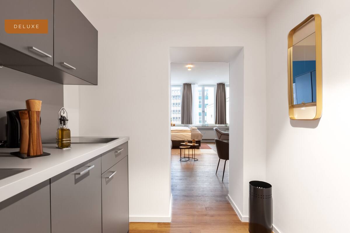 A modern kitchen with grey cabinets, a wooden countertop, and a black trash bin. A dining area with a table and chairs is visible through an open doorway.