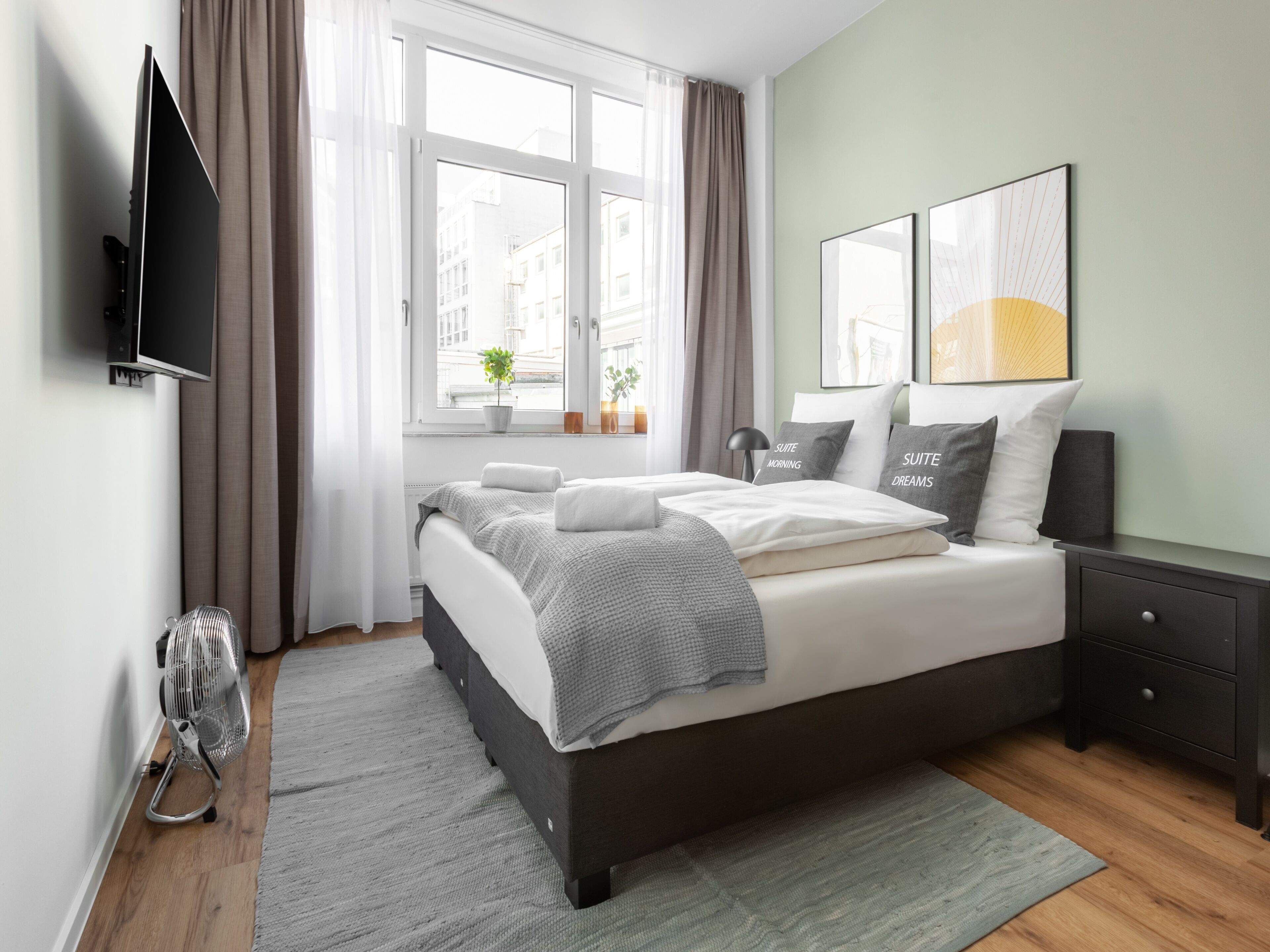 Un dormitorio moderno con una cama grande, un televisor de pantalla plana montado en la pared, una ventana con cortinas y dos cuadros enmarcados.