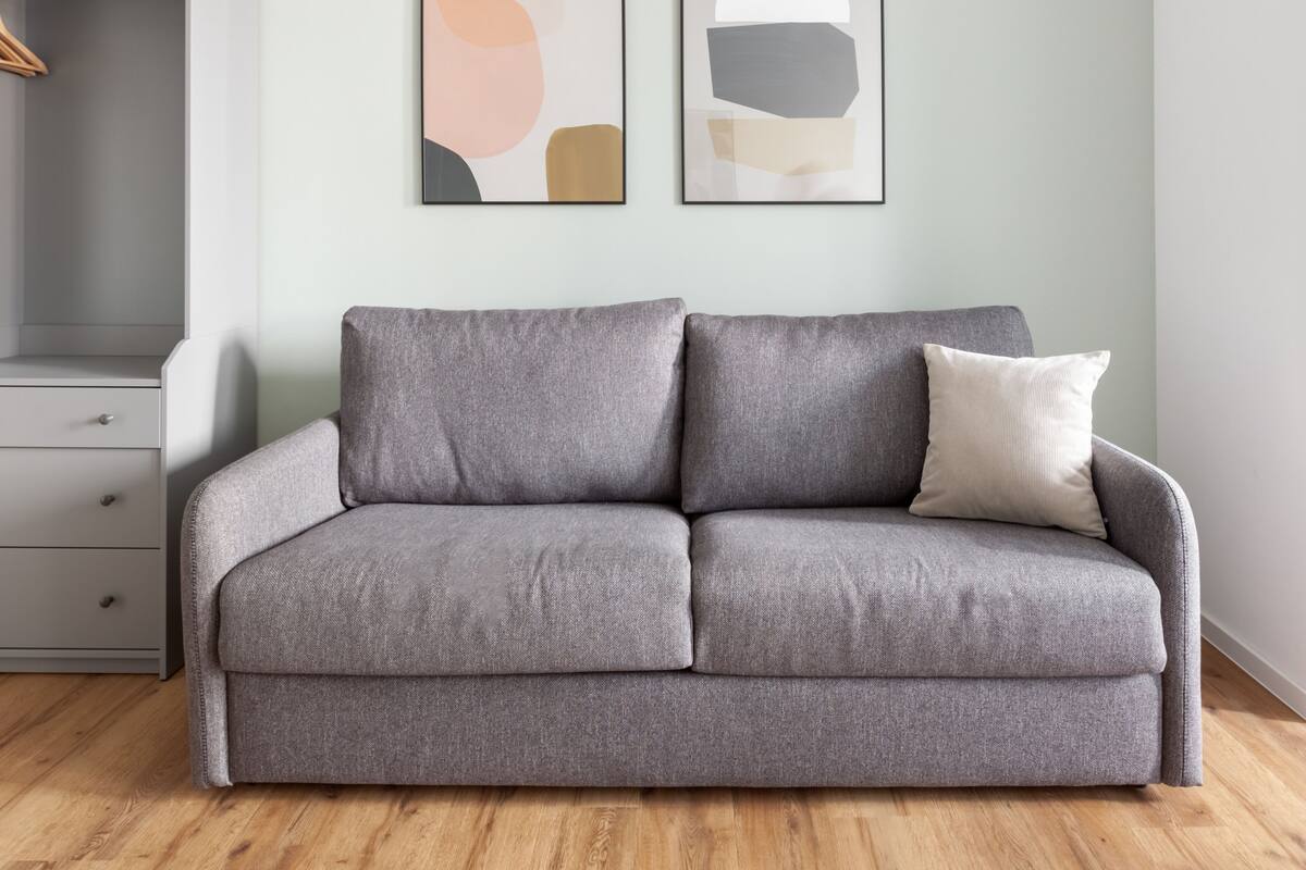 A grey sofa with a cushion, a wooden floor, a grey wardrobe, and two abstract paintings on the wall.