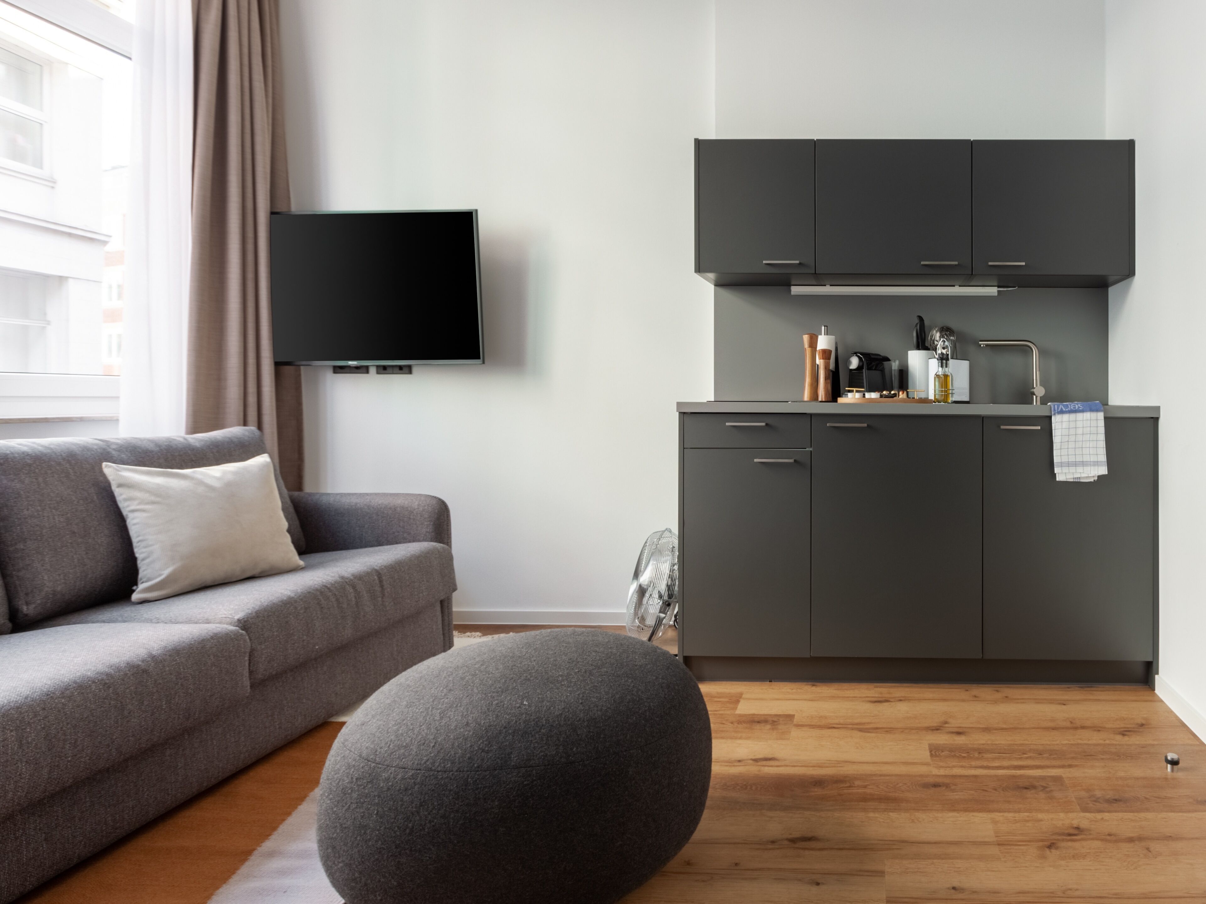 A modern living room with a grey sofa, a grey ottoman, and a flat-screen TV mounted on the wall.