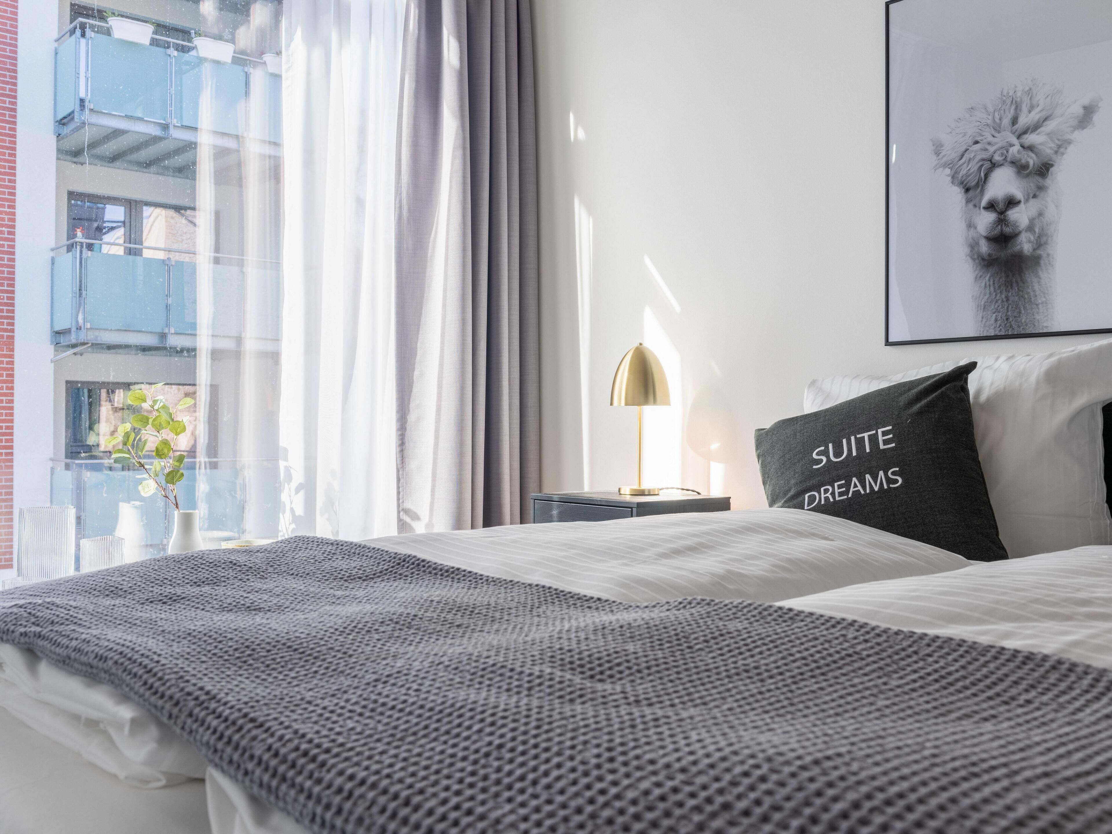 Een hotelkamer met een bed, een bedlamp, een kussen met tekst, een lijstje op de muur en uitzicht op het balkon.