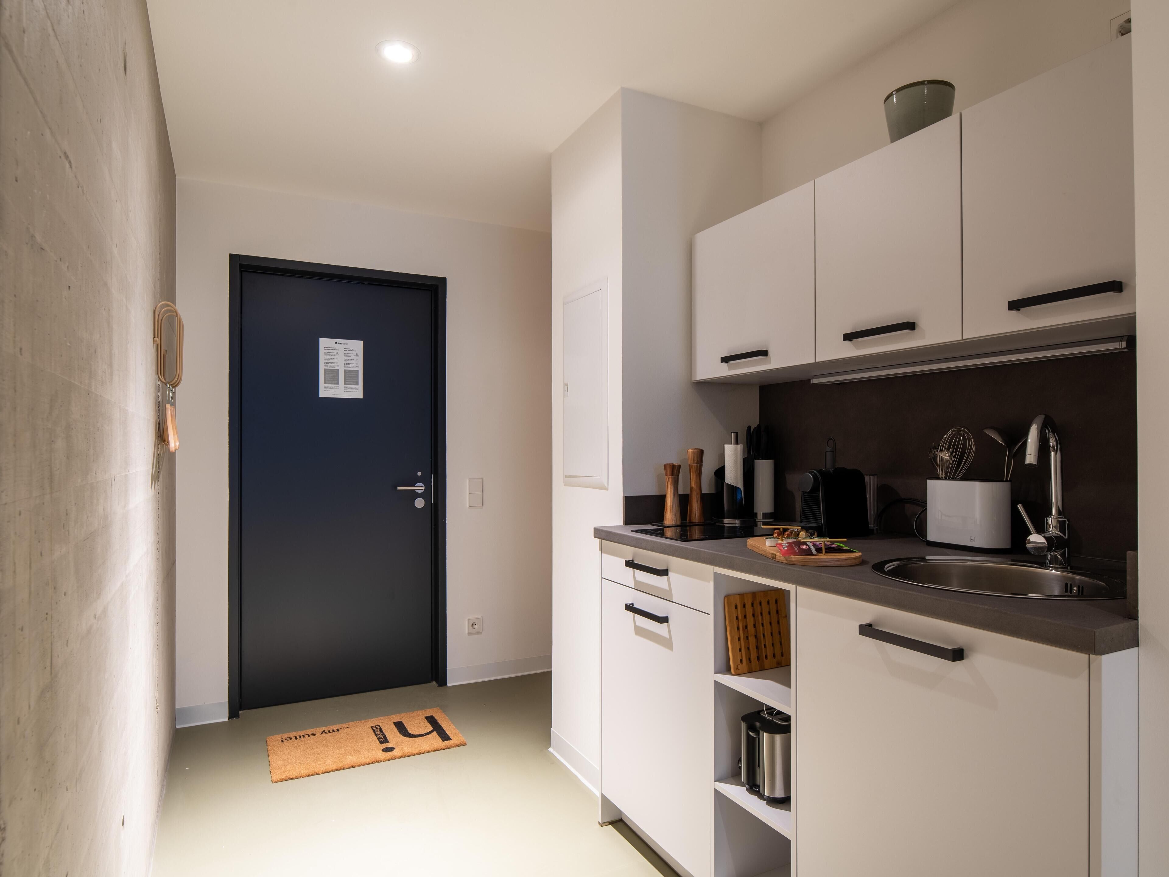 Une cuisine moderne avec des meubles blancs, une porte noire et un paillasson orné de la lettre « H ».