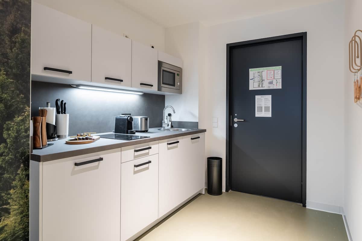 A modern kitchen with white cabinets, a microwave, a kettle, and a sink. There is a black trash bin and a door with a notice on it.