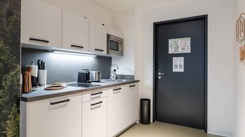 Cocina moderna con armarios blancos, microondas, hervidor, y un lavamanos. Hay un cubo de basura negro y una puerta con un aviso.