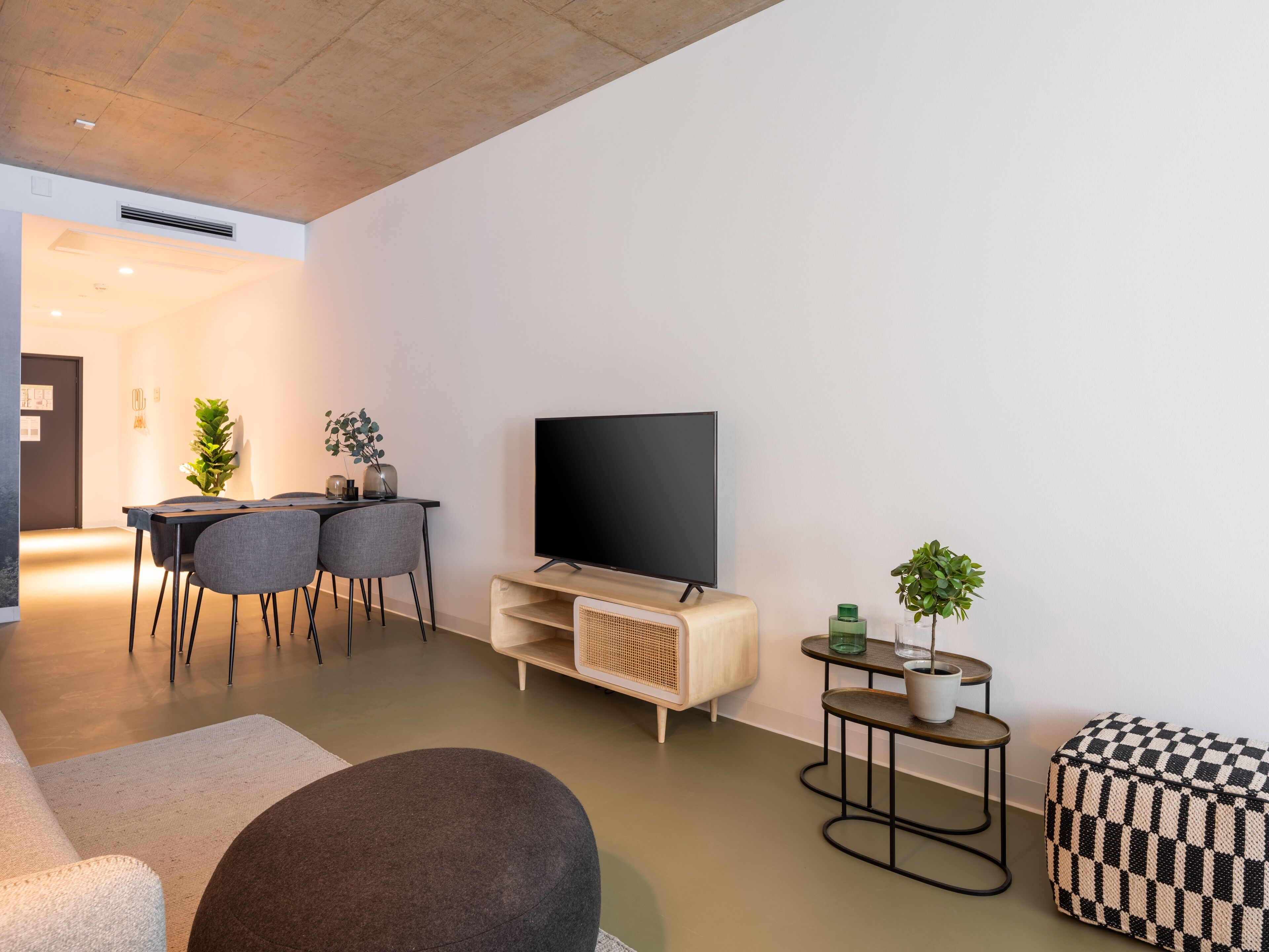 Una sala de estar moderna con televisor de pantalla plana, una mesa de comedor con sillas, un sofá, una mesita redonda con una planta y un reposapiés a cuadros.