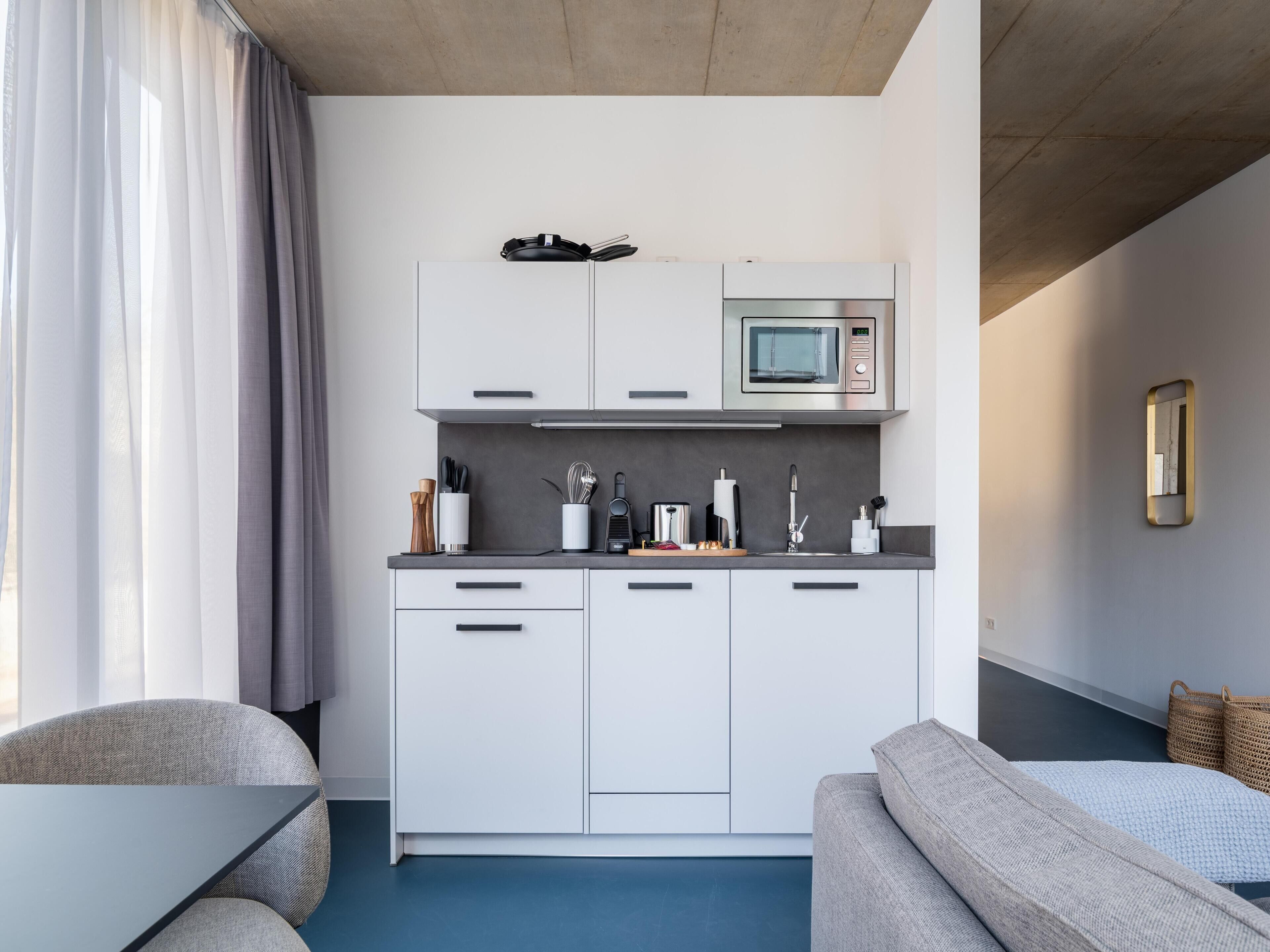 Een moderne keuken met witte kasten, een magnetron en diverse keukengerei. In de woonkamer staat een bank en een tafel.