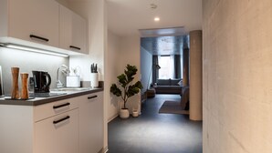A modern kitchen with white cabinets, a coffee machine, and a sink. There is a plant and a sofa in the adjacent living area. - Limehome Erfurt Maximilian-Welsch-Str. (Erfurt)