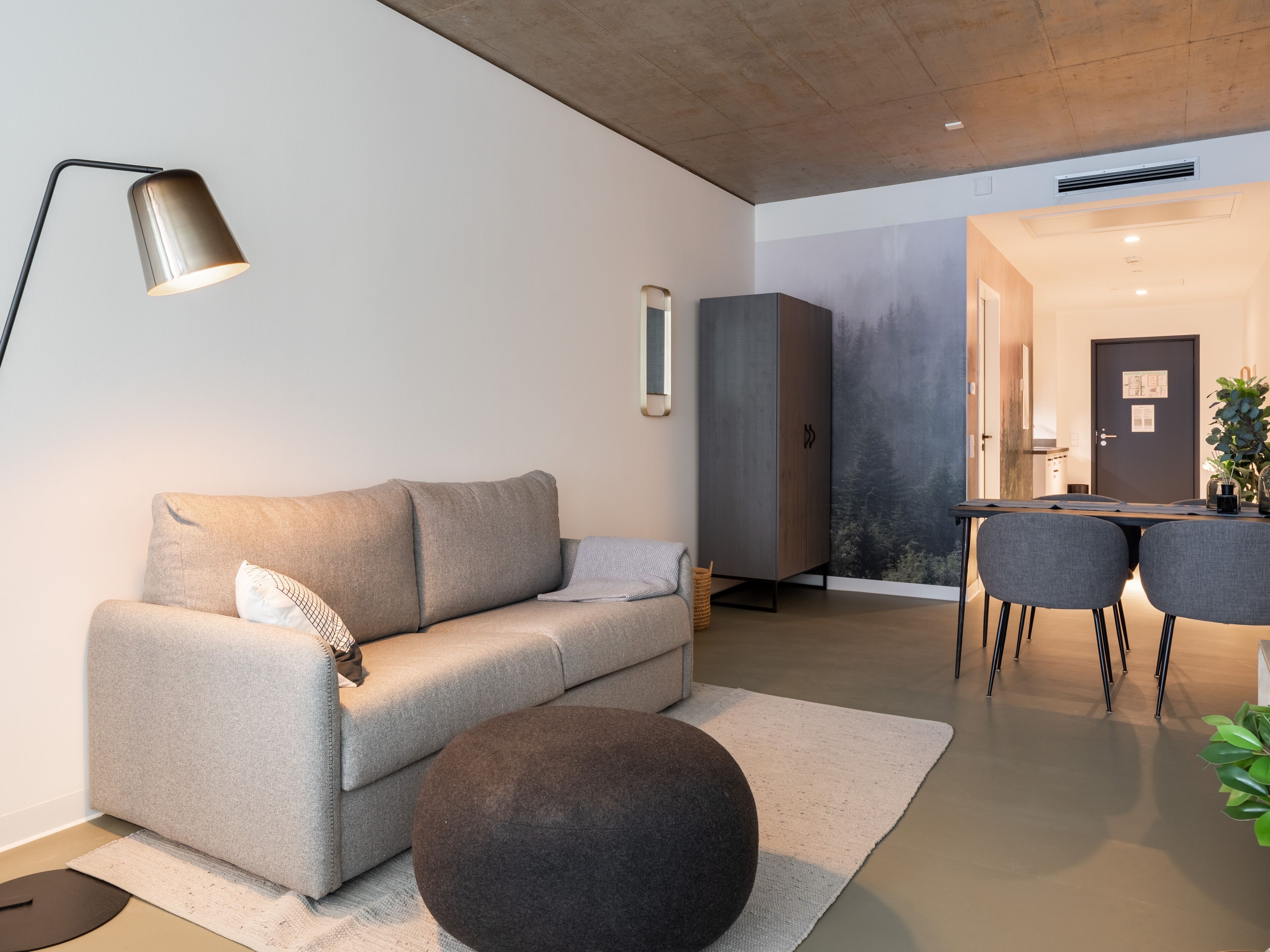 A modern living room with a grey sofa, a round ottoman, a floor lamp, and a dining area with two chairs and a table.