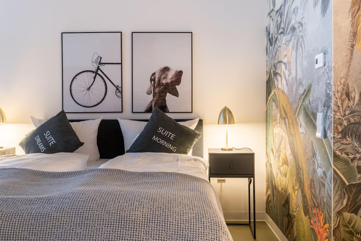 A neatly made bed with two pillows, a wall-mounted bicycle and dog picture, a nightstand with a lamp, and a tropical mural.