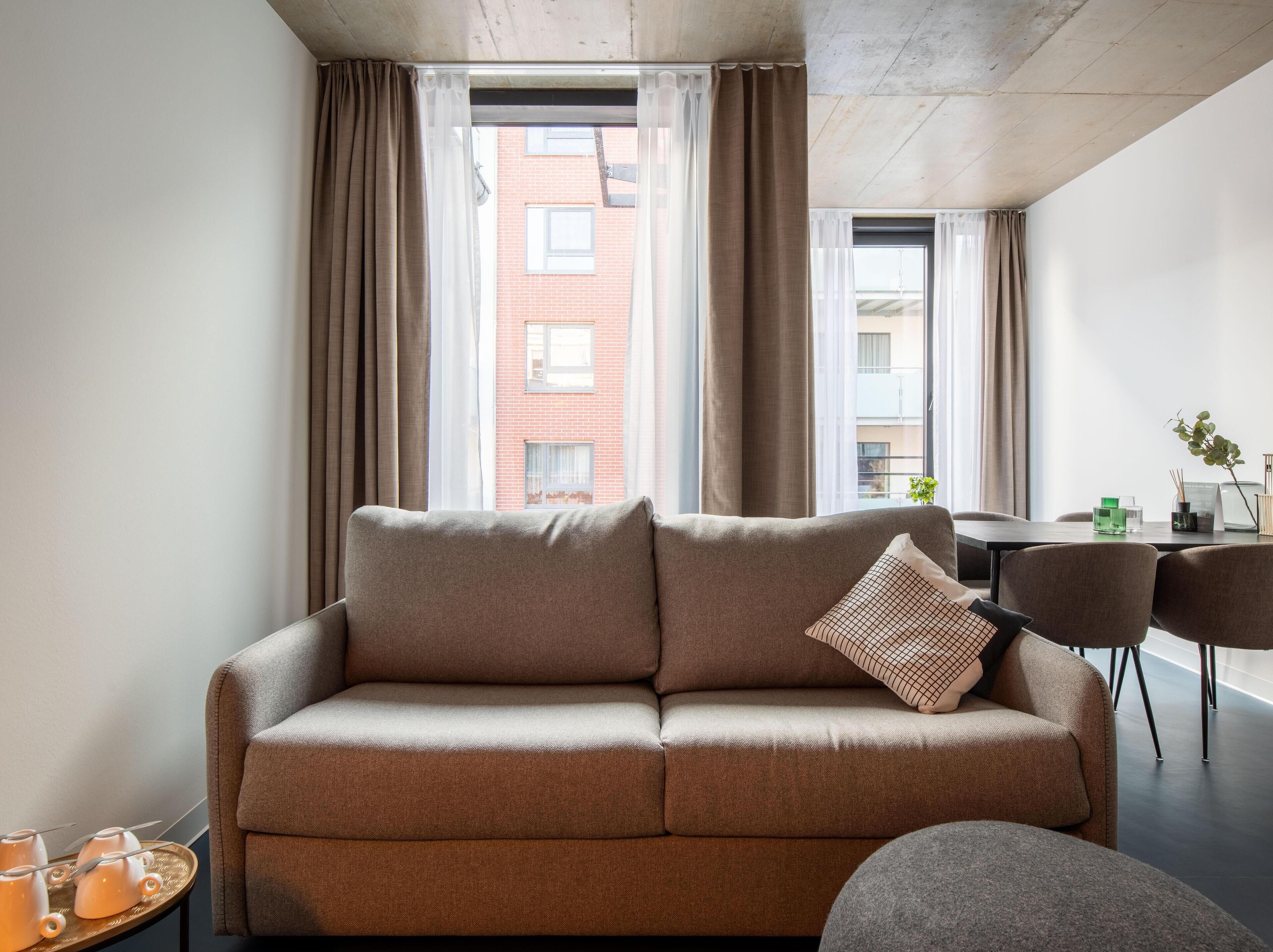 A modern living room with a sofa, a dining table, and chairs. There are curtains on the windows, and a view of a building outside.