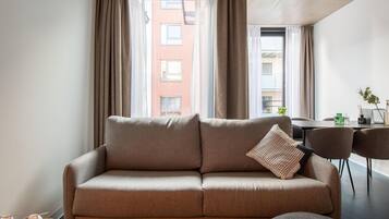 A modern living room with a sofa, a dining table, and chairs. There are curtains on the windows, and a view of a building outside.