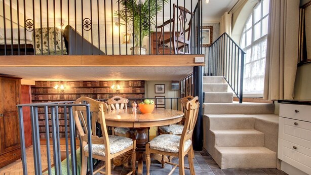 A dining area with a wooden table and chairs, a bookshelf, and a staircase leading to an upper level. - Carnegie Library Bronte Apartment (Swinton)