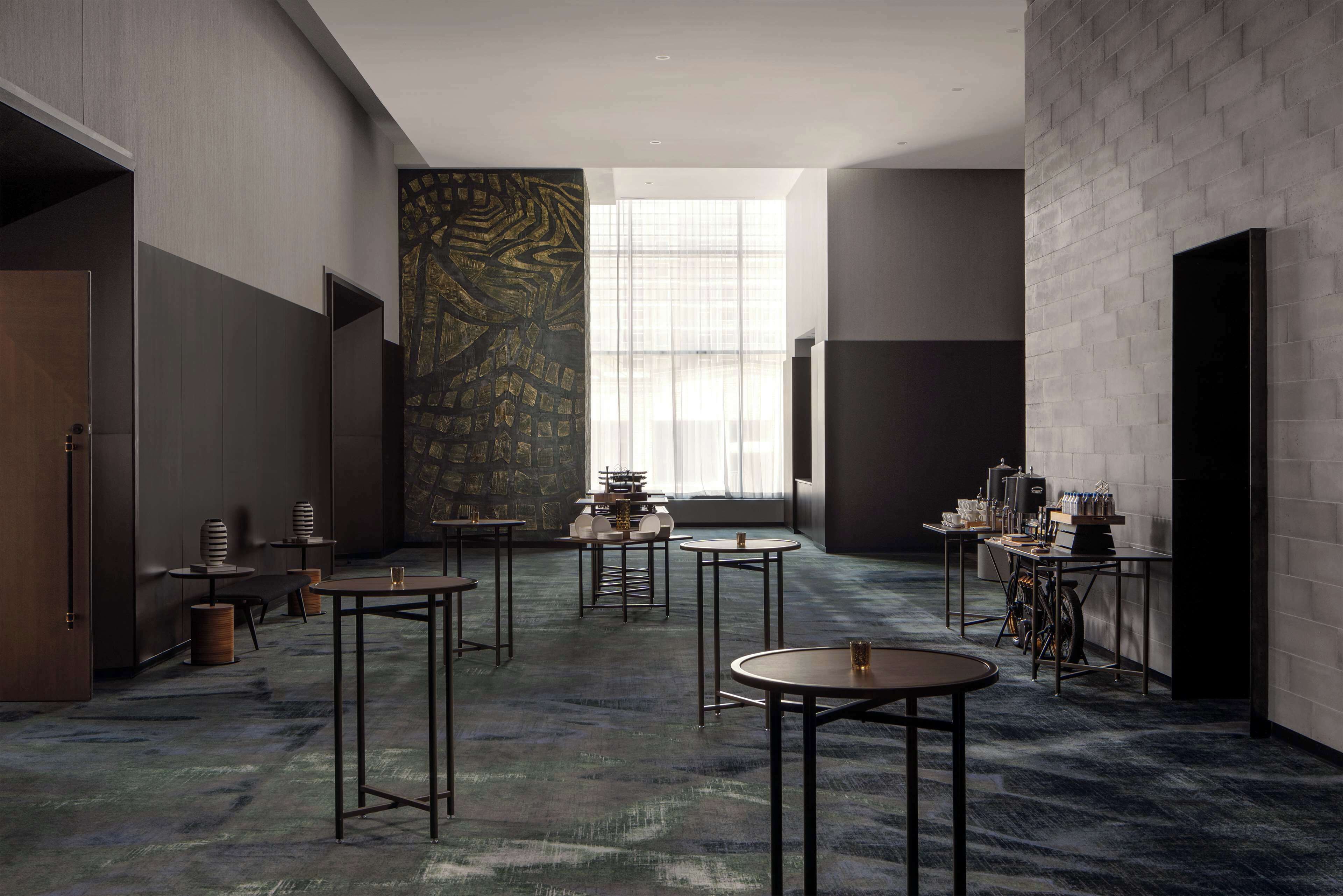 Un intérieur moderne avec un mélange de murs aux couleurs claires et foncées, un mur orné de motifs et un sol recouvert de moquette. On y trouve plusieurs tables et chaises disposées pour les repas ou les réunions.