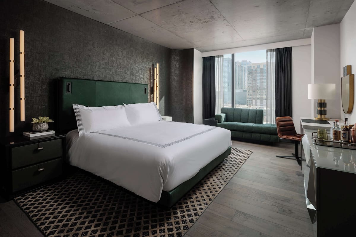 A modern hotel room with a large bed, a desk with a lamp, a chair, and a view of the city.