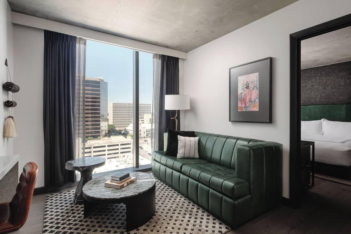 A modern hotel room with a green leather sofa, a round coffee table, a large window with curtains, a framed artwork on the wall, and a bed with white linens.