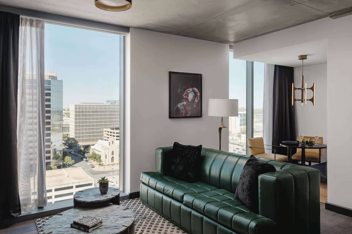 A modern hotel room with a green leather sofa, a small round table, a floor lamp, and a large window offering a city view.