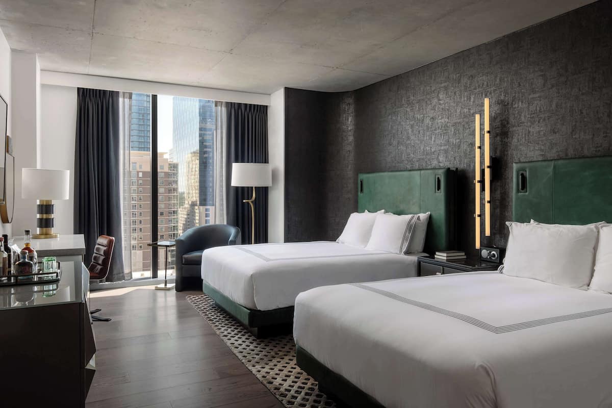 A hotel room with two beds, a desk with a lamp, a chair, and a view of the cityscape.