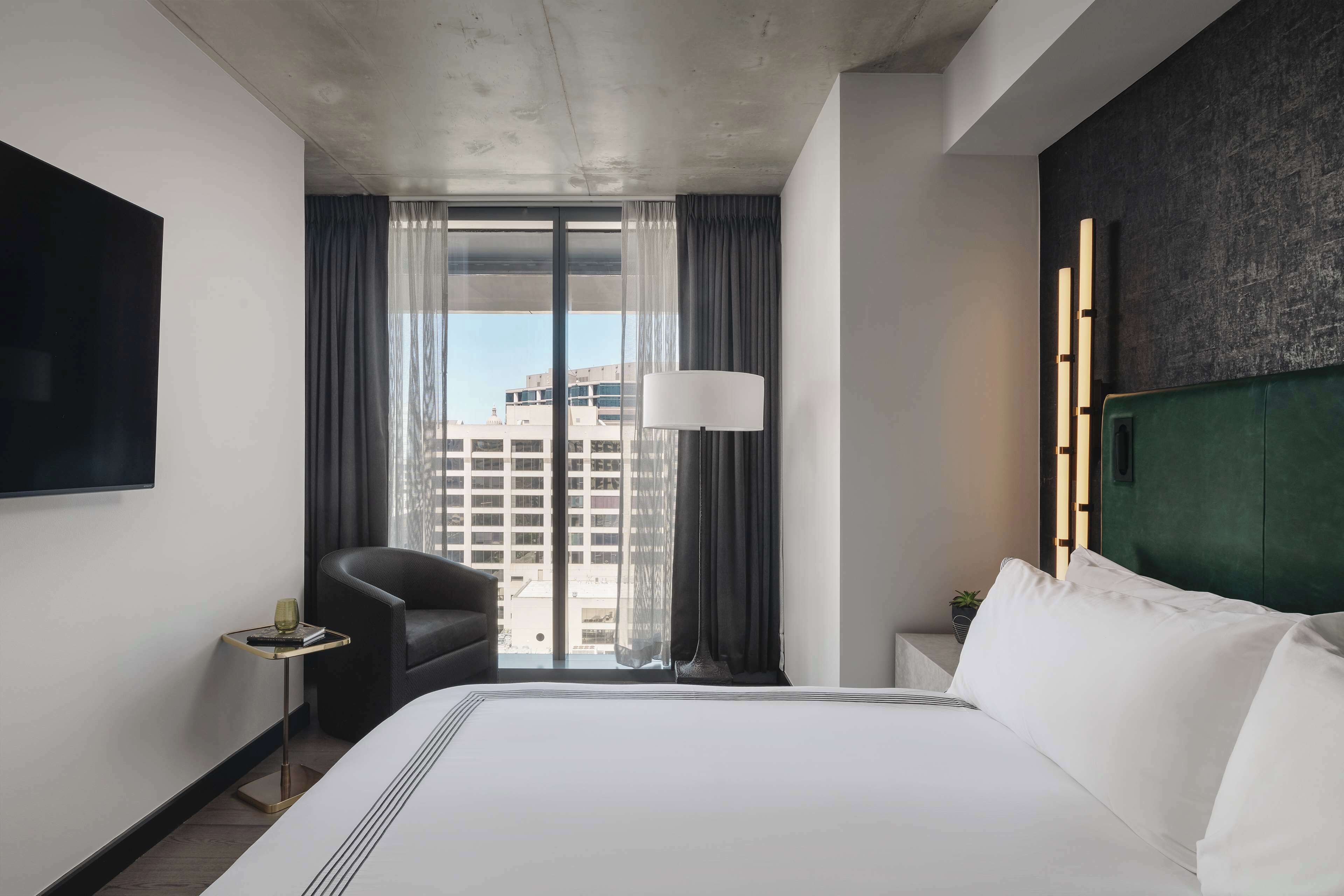 A hotel room with a large bed, a chair, a small table, a lamp, and a view of buildings through the window.