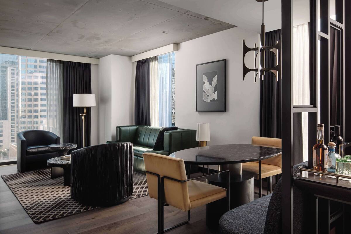 A modern hotel room with a green sofa, a black armchair, a round dining table, and a bar area with bottles.