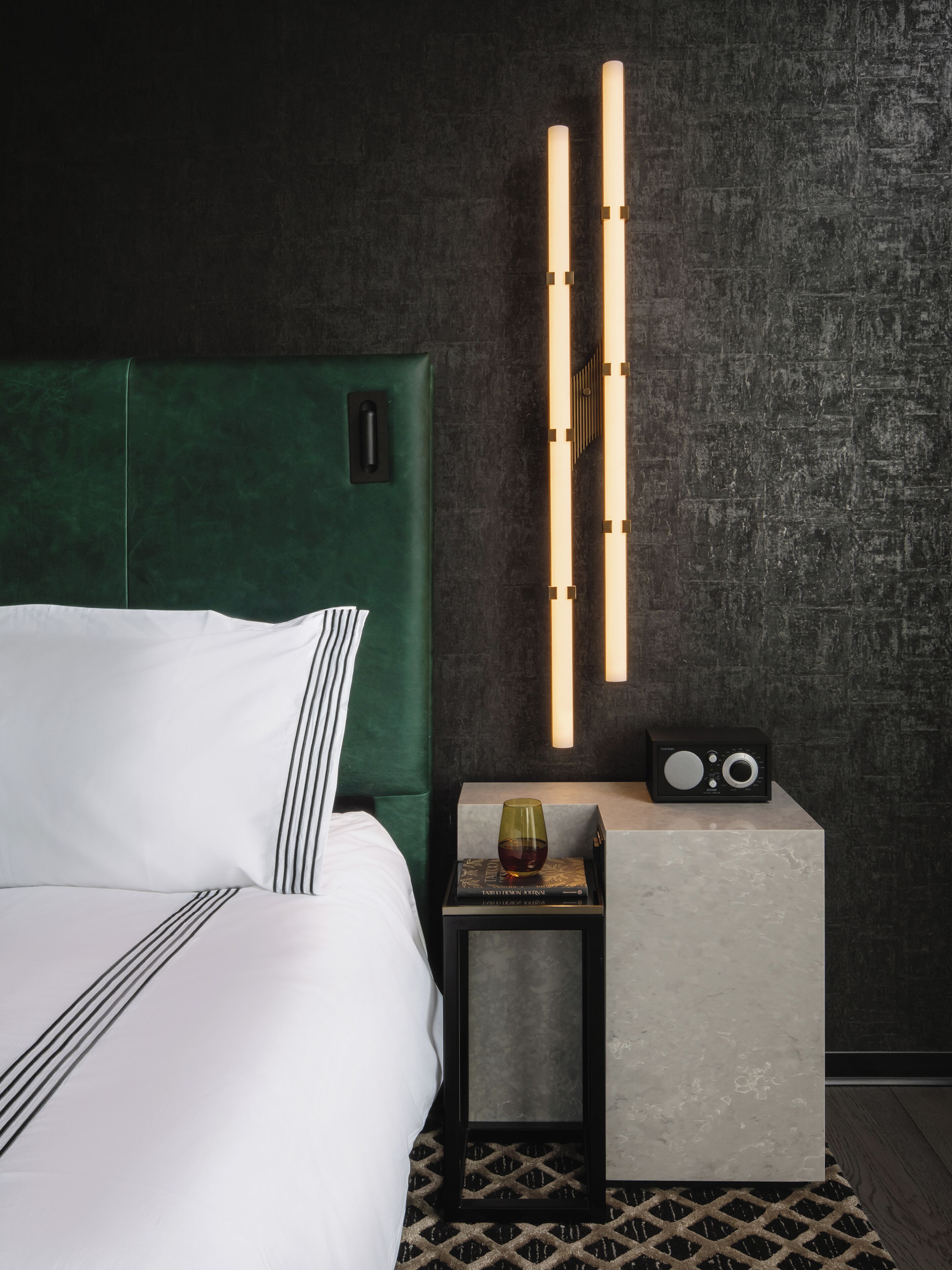 A neatly made bed with white and black striped bedding, a green upholstered headboard, a bedside table with a radio, and a wall-mounted light fixture.