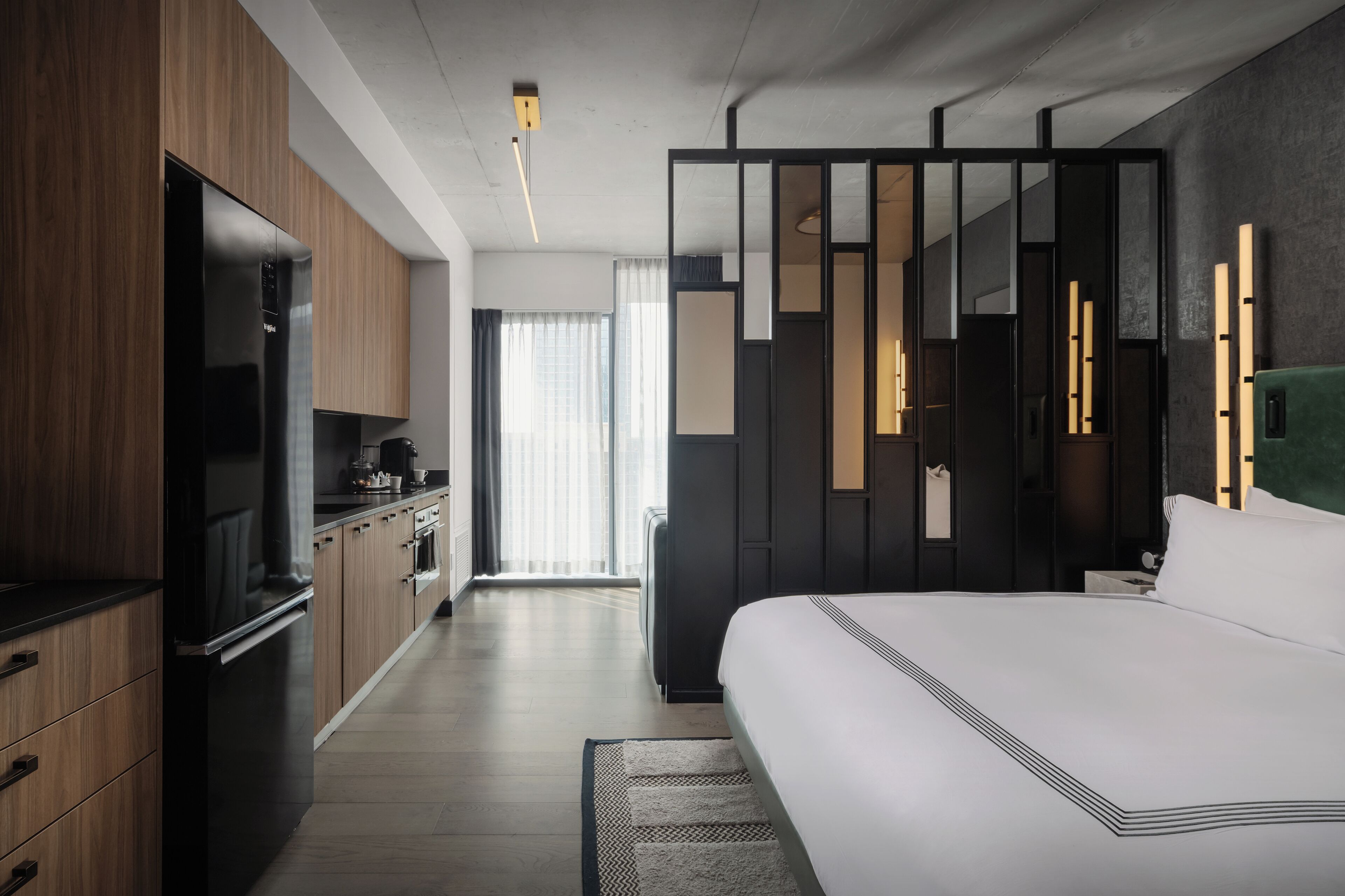 A modern hotel room with a large bed, a flat-screen TV, a kitchenette, and a sliding door leading to a balcony.