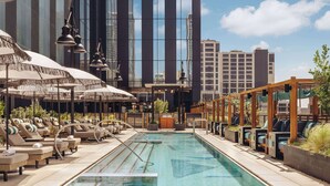 Una piscina en la azotea con sillones y sombrillas, rodeada de edificios altos.