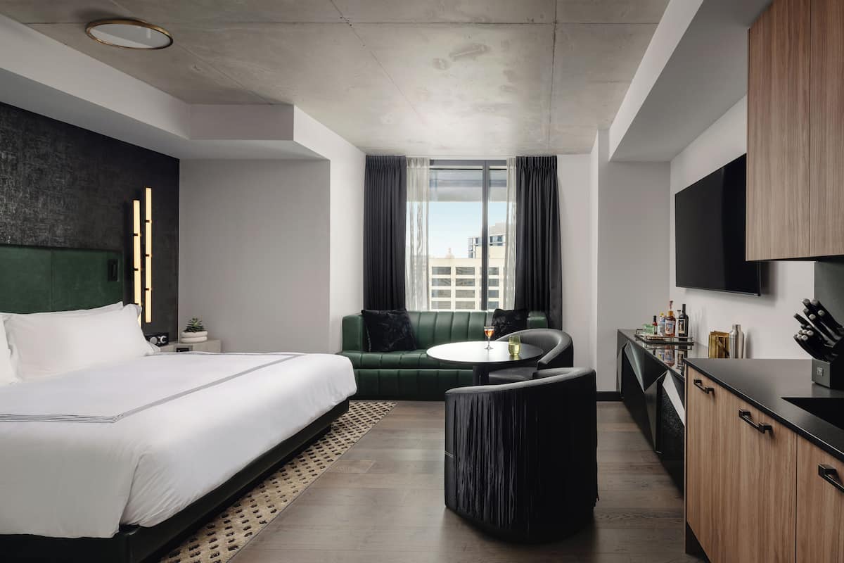 A modern hotel room with a bed, a green sofa, a small round table, a kitchen area, and a large window.