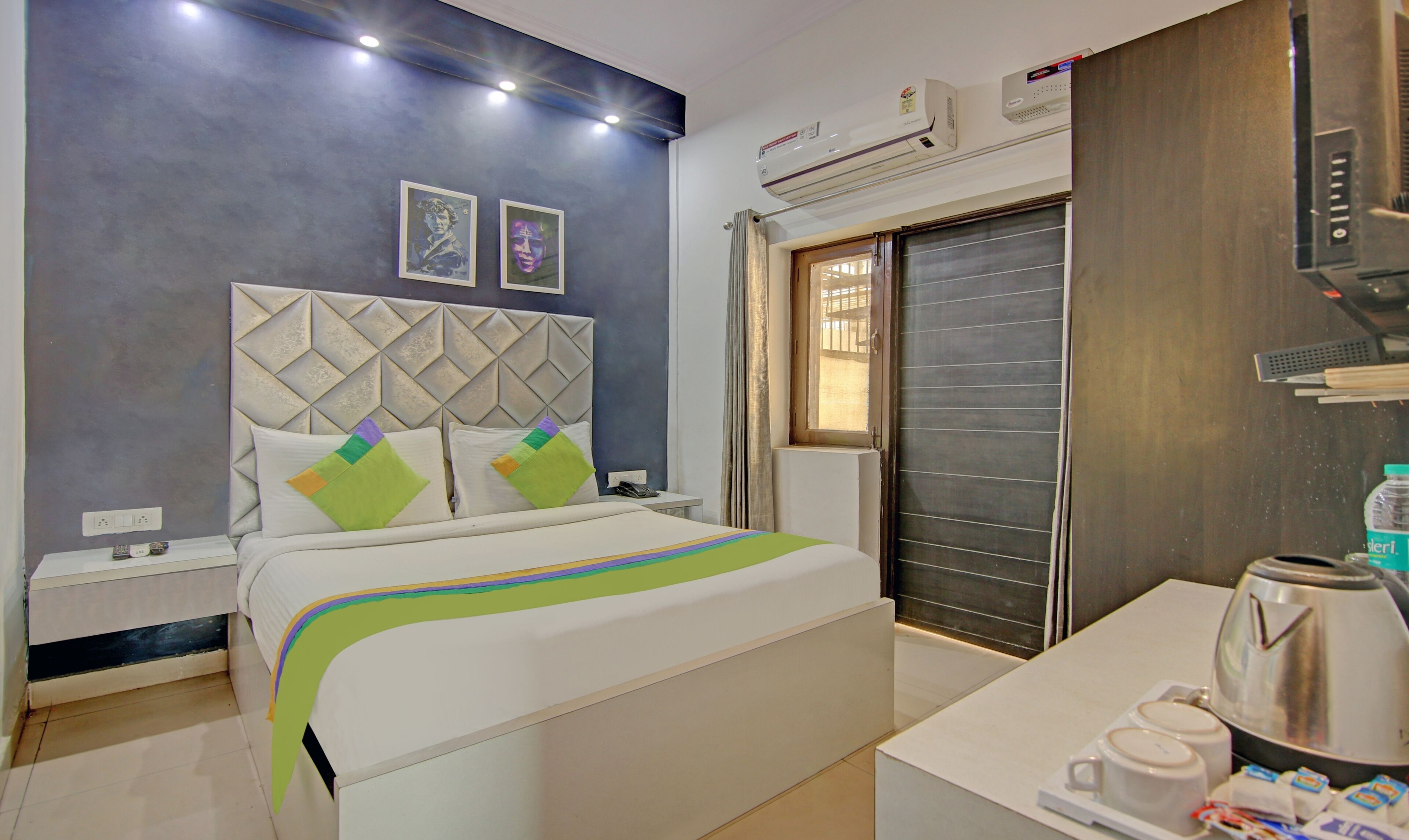 A hotel room with a bed, bedside tables, a desk with a kettle, and an air conditioning unit.