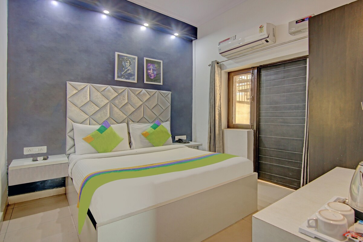 A hotel room with a bed, bedside tables, a desk with a kettle, and an air conditioning unit.