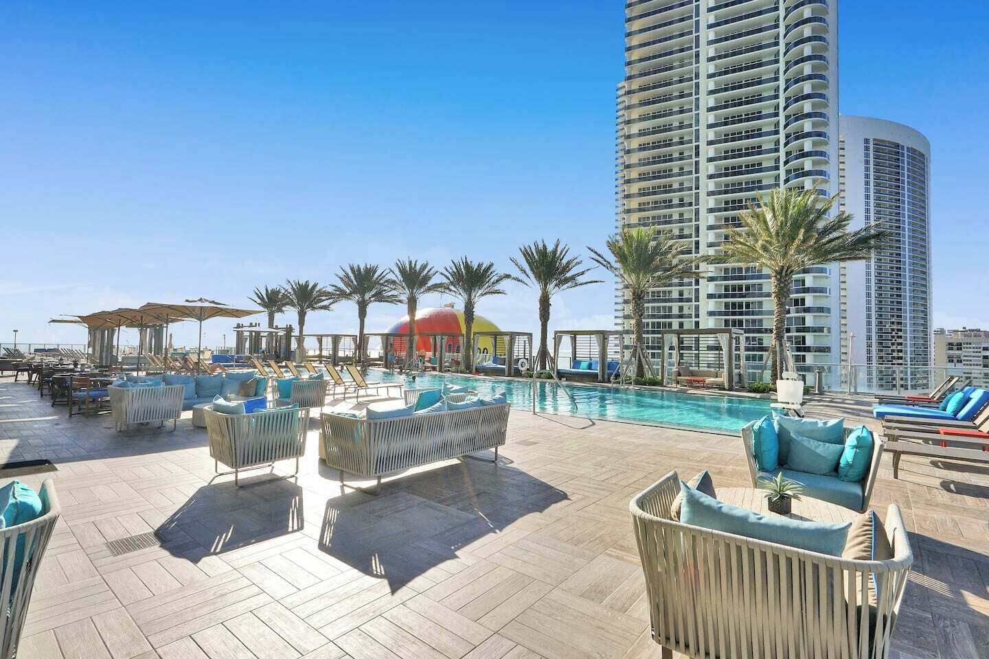 A rooftop pool area with lounge chairs and umbrellas, surrounded by palm trees and high-rise buildings.
