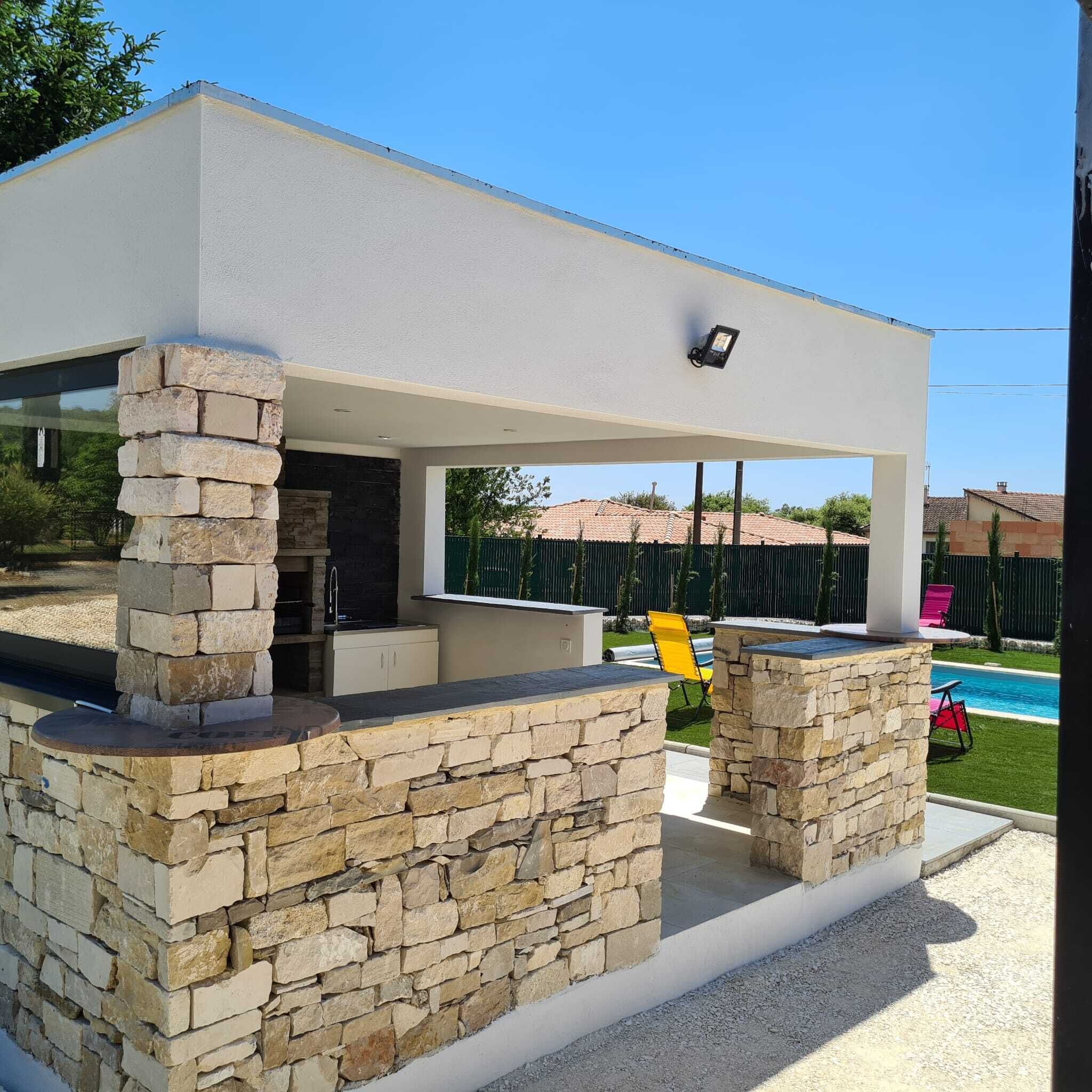 Un espace cuisine extérieur moderne, avec des piliers en pierre, un barbecue encastré et une piscine en arrière-plan.