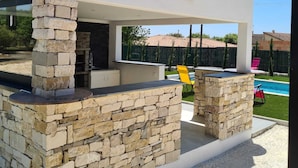Ein moderner Außenküchenbereich mit Steinpfeilern, einem integrierten Grill und einem Pool im Hintergrund.