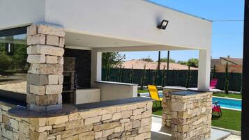 Un espace cuisine extérieur moderne, avec des piliers en pierre, un barbecue encastré et une piscine en arrière-plan.