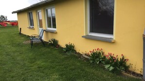 Un edificio amarillo con tres ventanas, una silla afuera y un jardín con tulipanes.