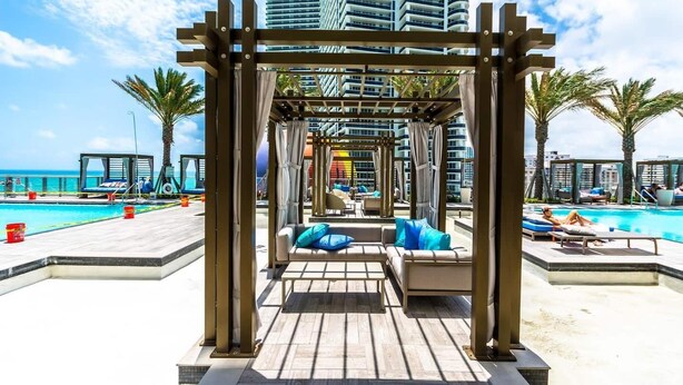 Un espace aménagé au bord de la piscine, avec une pergola en bois, des chaises longues et une vue sur l’océan et les immeubles de grande hauteur.