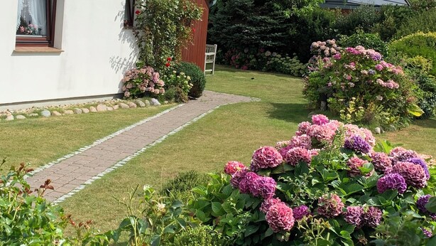 Una casa de dos pisos con fachada blanca y detalles marrones, un jardín cuidado con flores rosadas y un cielo azul despejado.