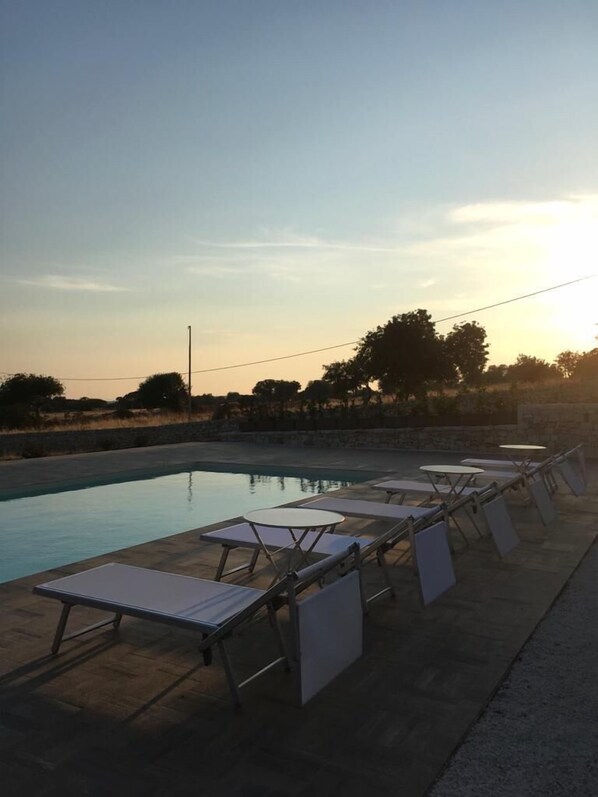 Un espace aménagé au bord de la piscine, avec des chaises longues et des tables, un mur en pierre et des arbres en arrière-plan.