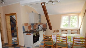 Une cuisine avec un mur recouvert de carreaux, des chaises en bois, une table à manger avec une nappe colorée et une fenêtre laissant entrer la lumière naturelle.