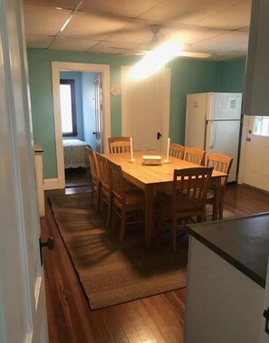 Zona pranzo con tavolo e sedie in legno, cucina con frigorifero e porta che conduce a una camera da letto.