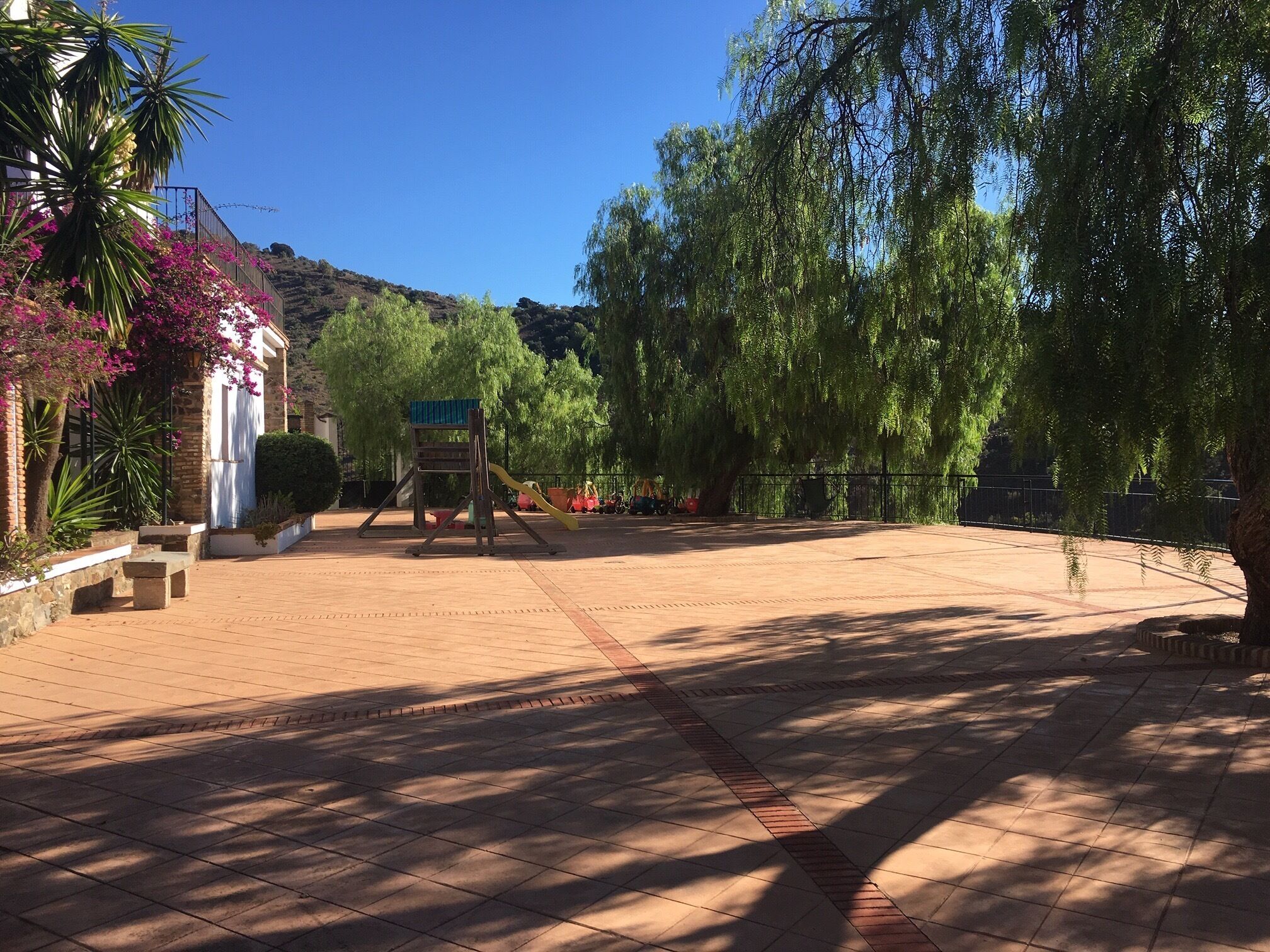 A paved courtyard with a playground, surrounded by trees and a building with a white facade.